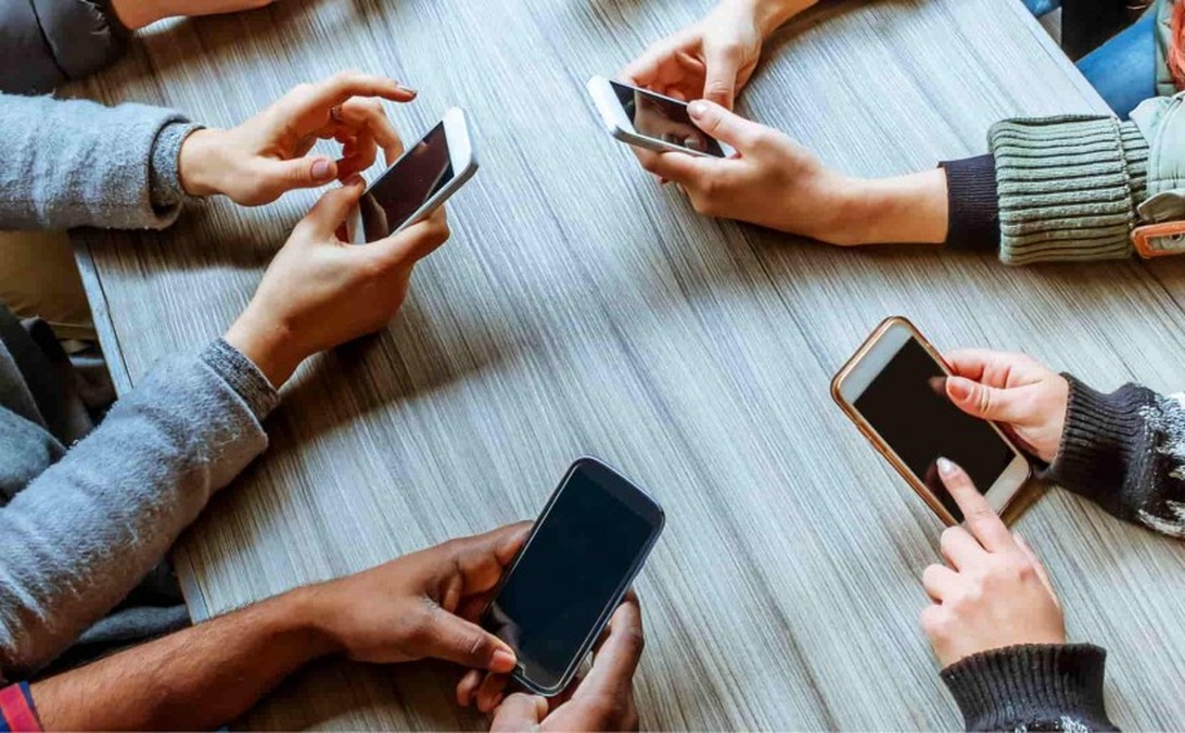 Flera händer som håller upp olika smartphones runt ett träbord.